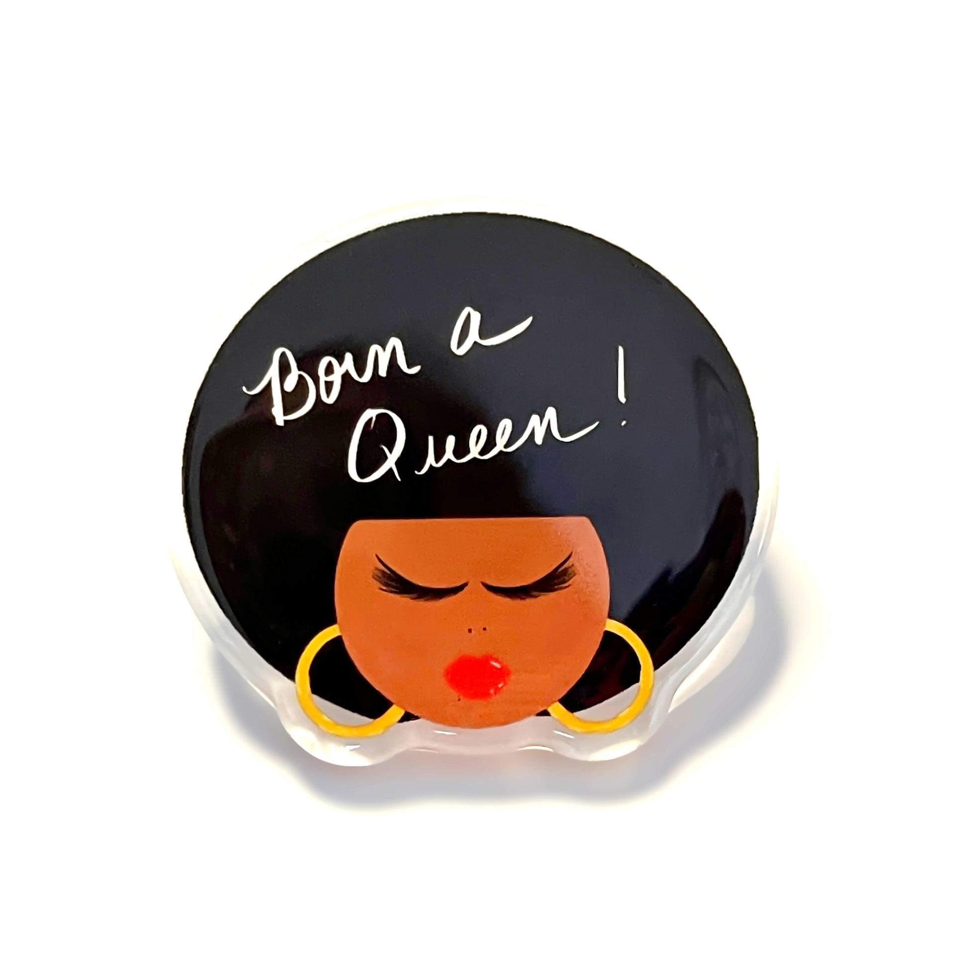 A round glossy black enamel pin featuring the text 'Born a Queen!' in white script and a stylized face with black hair, a brown skin tone, red lips, and yellow hoop earrings, displayed on a white background.